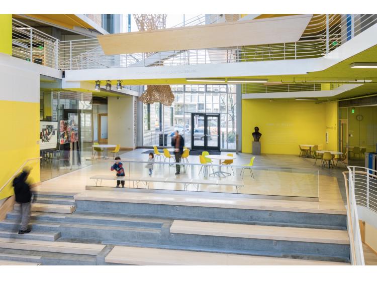 The atrium of Karl Miller Center, with people coming and going. On an elevated walkway, a line of white italic text reads, "how can we assemble ourselves..."
