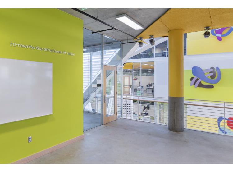 A green wall at left, and at right is a view into the atrium of Karl Miller Center. On the green wall is a line of raised white italic text reading, “to rewrite the structures of value.”