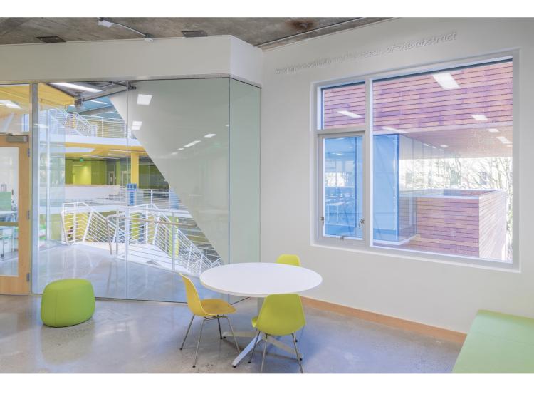 A seating area with white walls and large windows. High on the wall at right is a line of text in a raised white italic font. It reads, “to share a solidarity in the ocean of the abstract.”