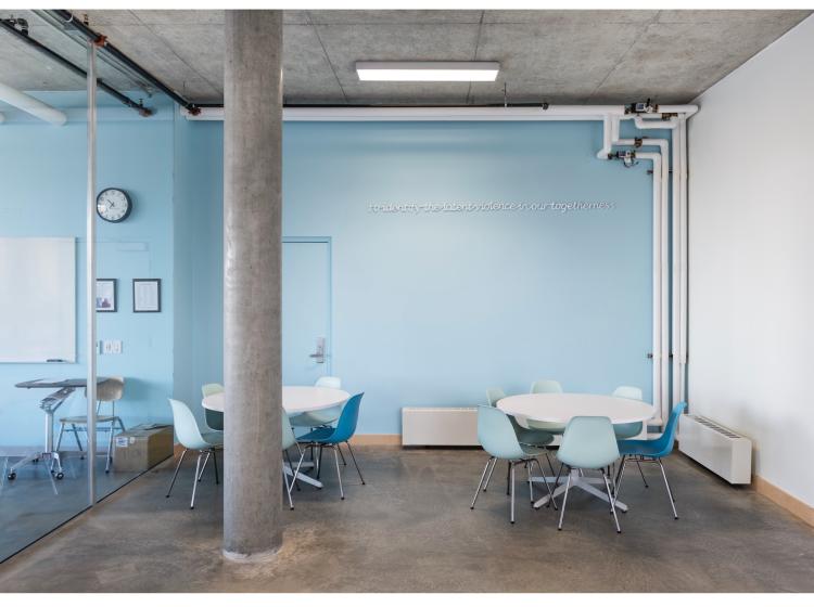 A seating area with light blue walls. High on the wall facing the viewer, a line of text appears, in a raised white italic font. It reads, “to identify the latent violence of our togetherness.”