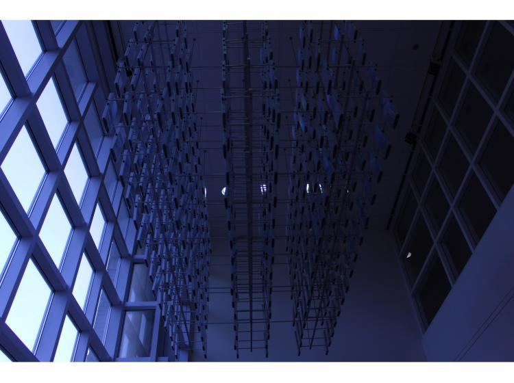 View from the side, looking up at the artwork in near darkness. Light is visible through windows at left, but the room and sculpture are only dimly lit.