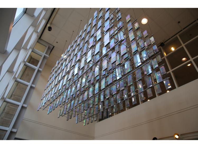 Suspended from the ceiling and lit by windows at left is a sculptural installation that consists of reflective rectangular elements hanging in a three-dimensional grid pattern.