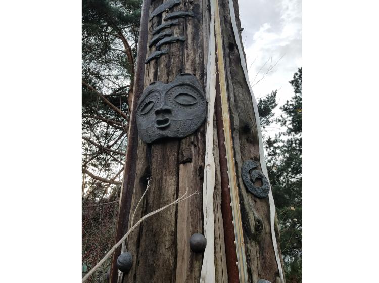Close-up of a section of the pole. The wood is roughly textured and decorated with small sculptural elementsm. Featured here is an abstracted metal face.