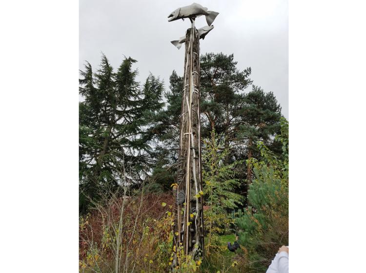 A tall wooden pole towers above tall grass, with trees in the background. The pole is decorated with sculptural elements in the shape of salmon and faces.