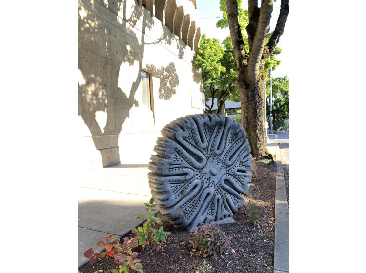 A granite sculpture sits in the dirt swale next to the sidewalk on 6th Avenue. The sculpture is shaped like a microscopic organism, somewhat resembling a wheel.