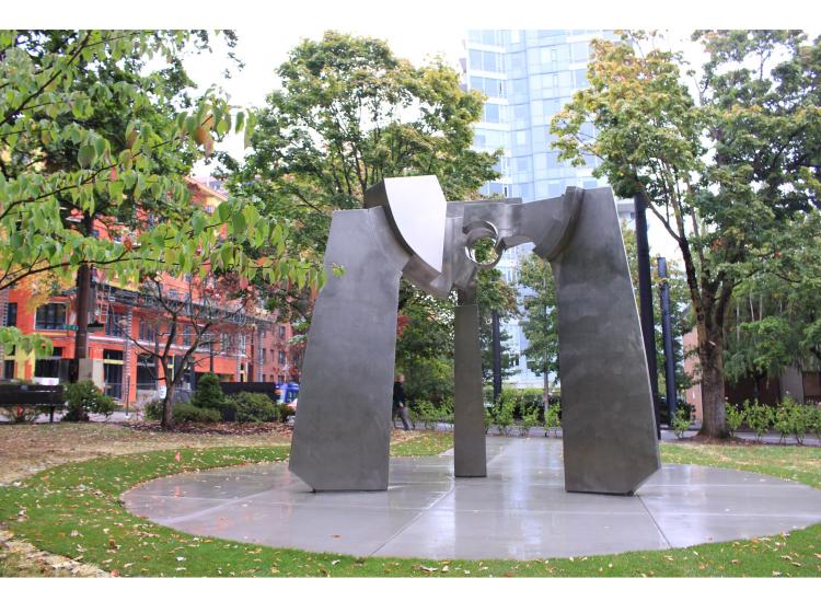 A large abstract steel sculpture on a round granite base surrounded by grass, buildings and trees. The sculpture has three “legs” meeting at the top in a joined area with a round hole through it.