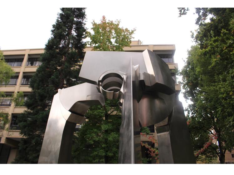 Close-up of the upper portion of the sculpture, with treetops and Science Building 1 in the background.