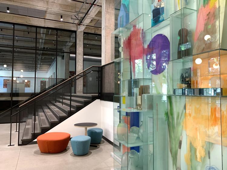 A large glass artwork is seen at right, which resembles a cluster of stacked glass display cases with colorful elements inside. At left is a flight of stairs.