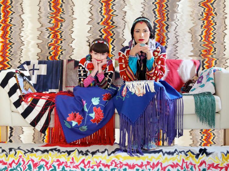 Vividly colored photo of a woman and adolescent girl sitting together on a sofa, looking at the viewer. They are adorned and surrounded in colorful Native American patterned textiles.