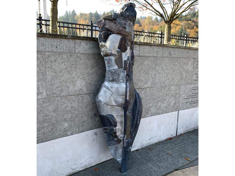 A torso-shaped abstract metal sculpture is seen in front of a low granite wall, with trees and hills visible in the background.