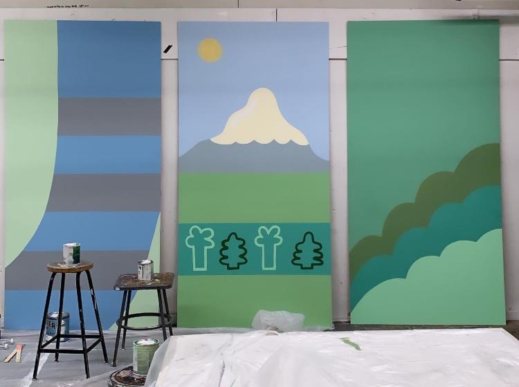 Pieces of the mural are seen in the process of being painted, leaning against the wall. In front of them are stools with cans of paint on them.