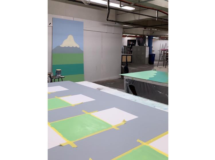 Pieces of the mural are seen in the process of being painted. In the foreground is a section with squares outlined by painter's tape.