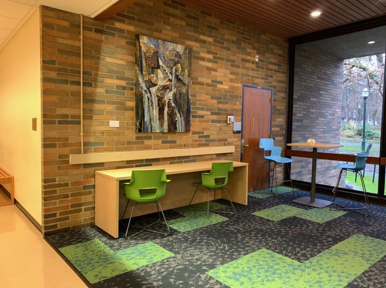 The painting is shown hung on a brick wall in a small study lounge, with windows to the right. In front of the painting is a small table with green chairs.