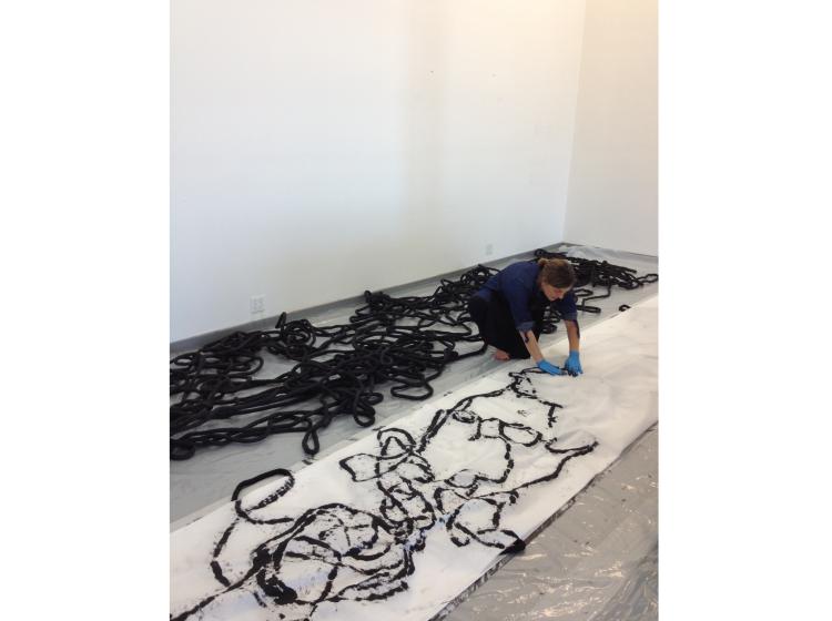 The artist is shown dyeing the rope-like cord black. Behind her on the floor is a mass of dyed black cord, and she is kneeling and pressing the wet dye out of a batch of cord using white paper