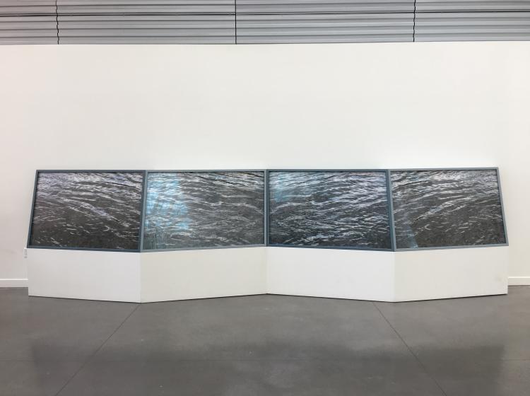 A long, low pedestal is seen against a white wall and shiny grey floor. The pedestal contains four forward-facing black and white photos of rippling water.