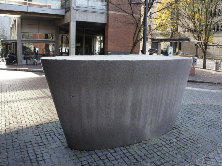 A large, abstract granite sculpture sits on the Urban Plaza. It is grey, shaped like a cross section of an inverted cone, and has a rough top surface and smooth sides.
