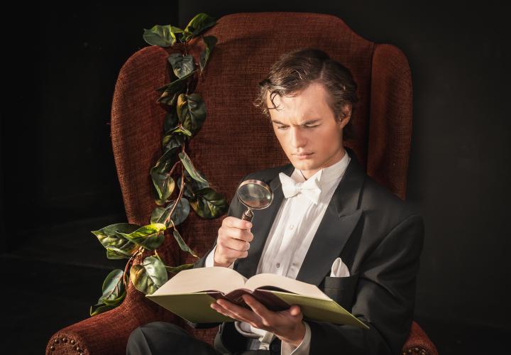 PSU Opera's Sherlock Holmes reading a book with a magnifying glass.