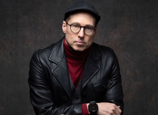 A man in a red turtle neck, black leather jacket, newsboy cap, and glasses.