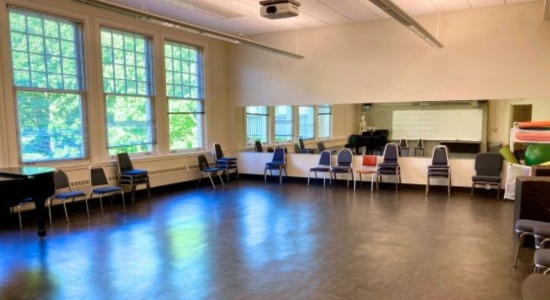 Large, mostly empty Theater rehearsal room in Lincoln Hall 319 with full wall mirror, large whiteboard, projector, and large windows the length of one wall looking out a tree tops.