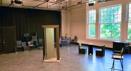 A large theater rehearsal room with lighting grid on tall ceilling, various set pieces like a theatrical door and furniture, and large windows the length of one wall facing tree tops.