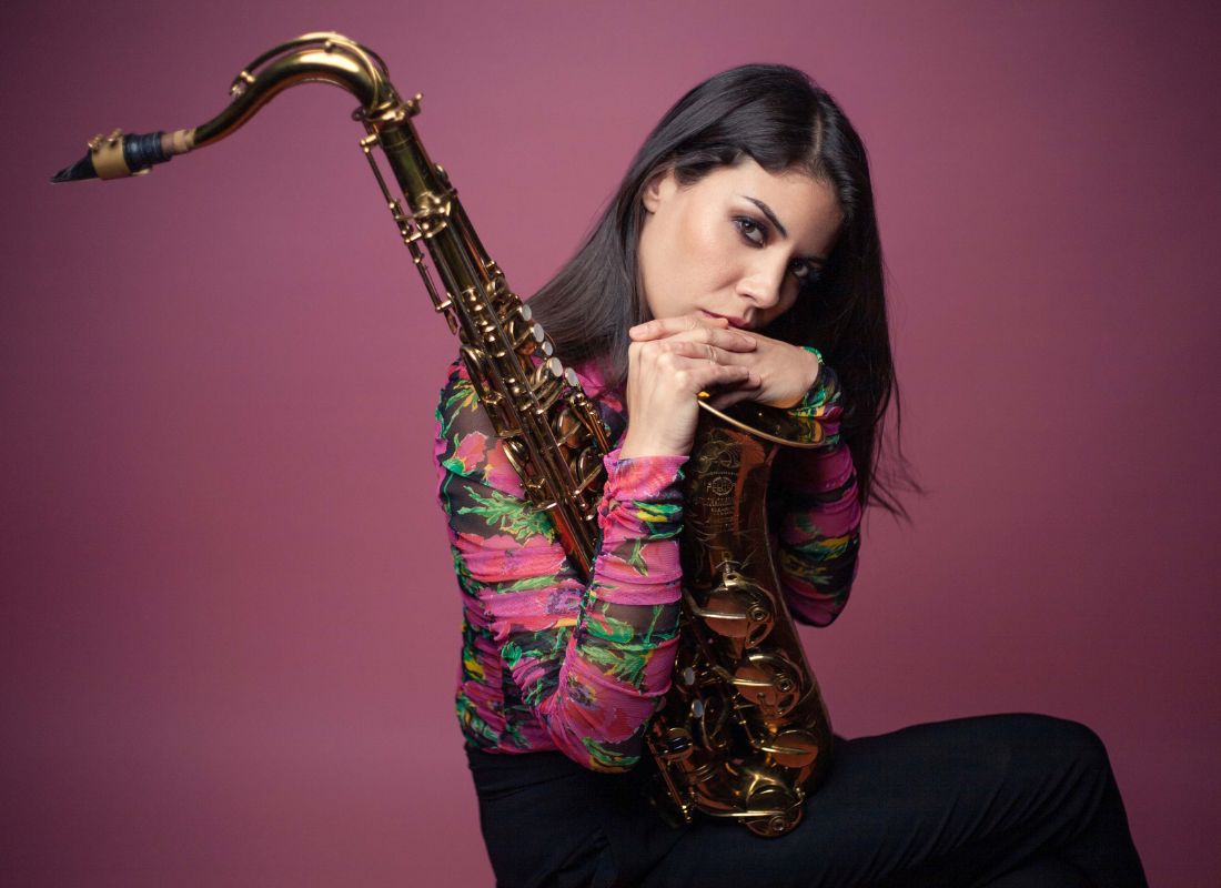 A woman looking seriously as she holds and leans on her saxohpone