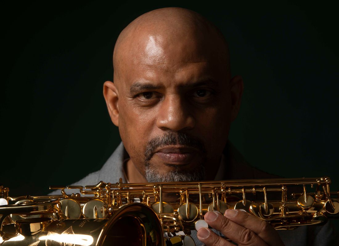 A man's face half drenched in shadow as he presents his saxophone forward.