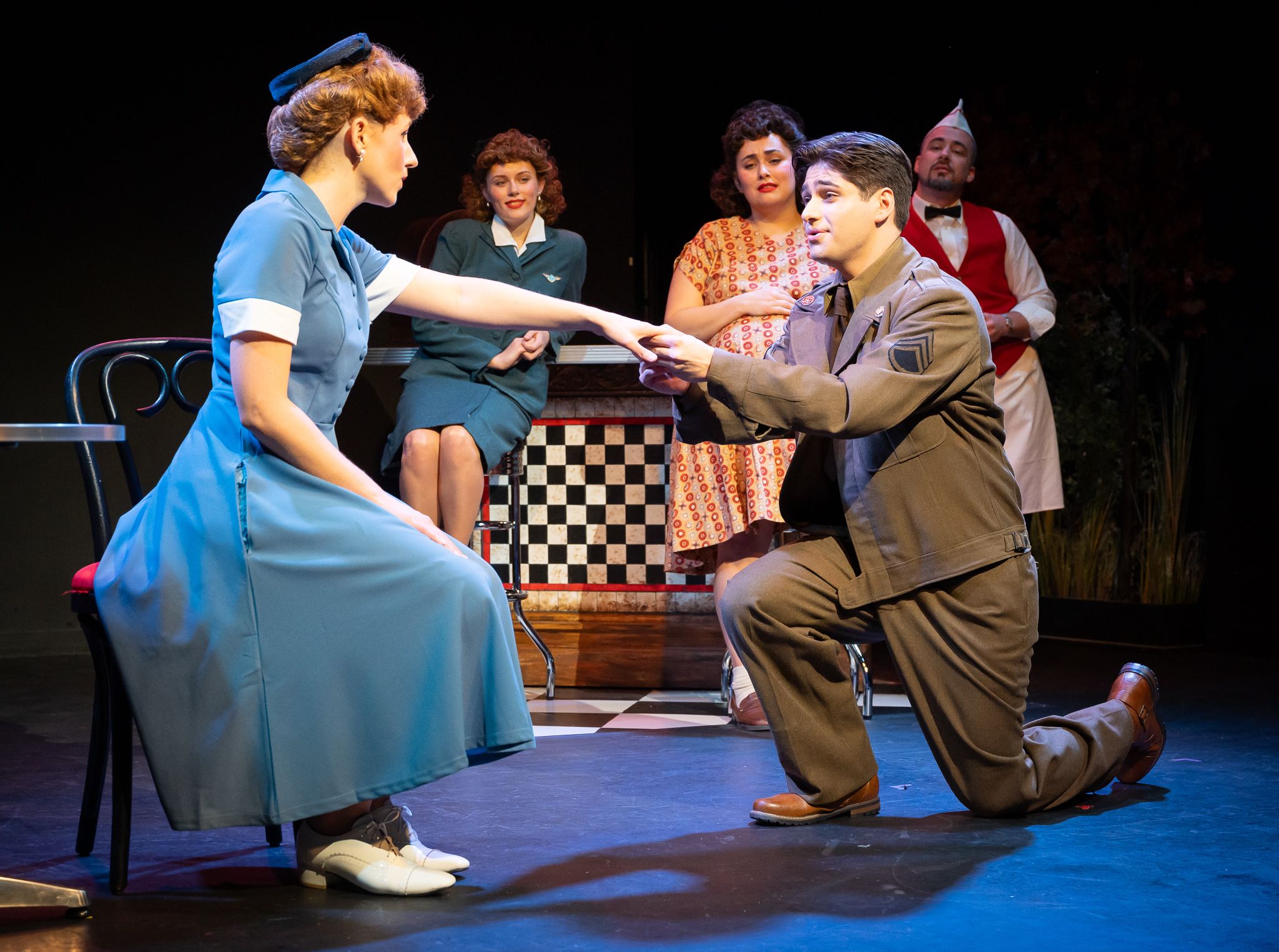 A woman in a dress sits in a chair as an American soldier kneels in front of her holding her hand. They are on stage in a 40s style diner with other characters watching them