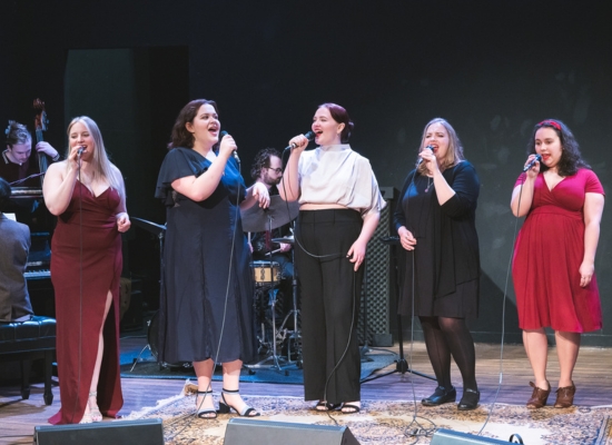 Five PSU Vocal Collective students singing on stage with a jazz band playing behind them.