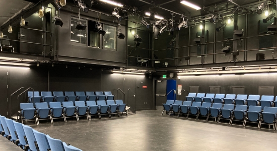 Lincoln Hall room 55 with blue audience chairs arranged along three of  the walls in two rows each wall, a sound booth and lighting grid above.