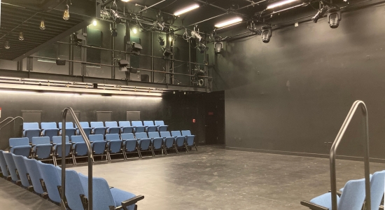 Lincoln Hall room 55 with blue audience chairs arranged along three of the walls facing the fourth blank wall, with two rows of seats on each wall, and a sound booth and lighting grid above.