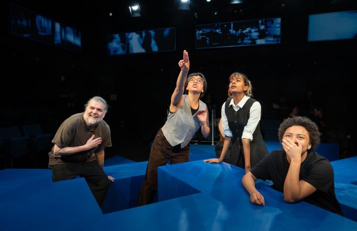PSU cast perform in Caryl Churchill's "Love and Information." four people are looking up at a TV screen with varying reactions. One if pointing.