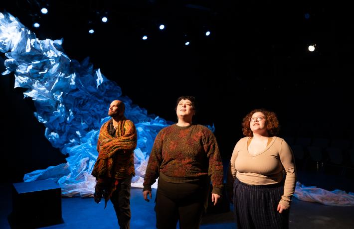 Scene from PSU Theater Arts' 2024 production of "Y/OURS." Three people stand looking up towards the light with a huge mountain of crumpled paper behind them.