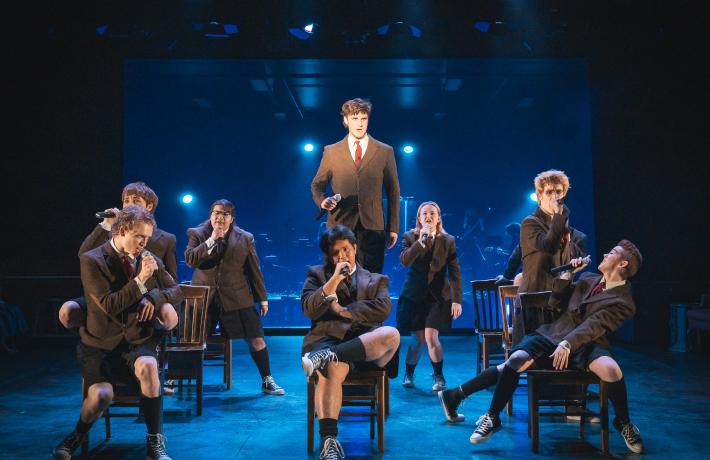 Young men in boarding school uniforms rocking out on classroom chairs with dramatic lighting.