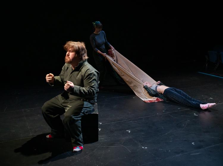 Actor pretending to drive a car while another actor drags a body away on a cloth. From PSU's Theater production of "Y/OURS."