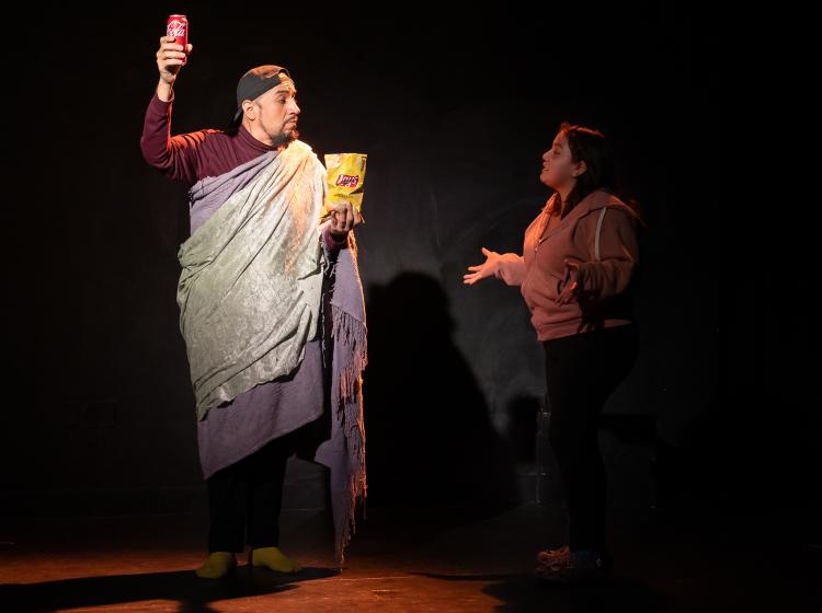 An actor standing like the Statue of Liberty with Coke instead of a torch and chips instead of  a book, and another actor talking to him. From PSU's Theater production of "Y/OURS."