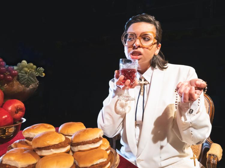 Image of PSU Theater Arts' Tartuffe drinking wine, holding a gold cross and face a table overflowing with burgers and other food.