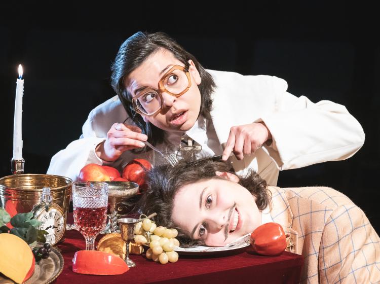 Image of PSU Theater Arts' Tartuffe looking suspicious as he cuts into Orgon's smiling head on a platter next to a table overflowing with food and wine.