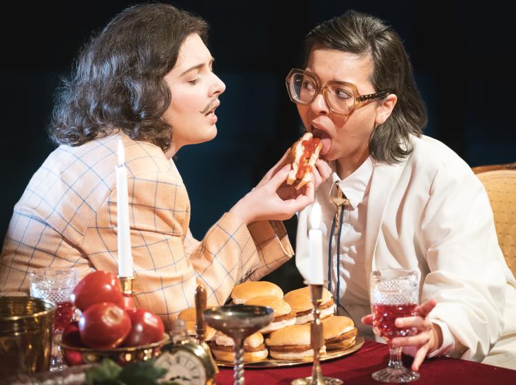 Image of PSU Theater Arts' Tartuffe is being fed a cheese burger by Orgon as they sit in front of a banquet table full of wine, fruit, cheese and candles.
