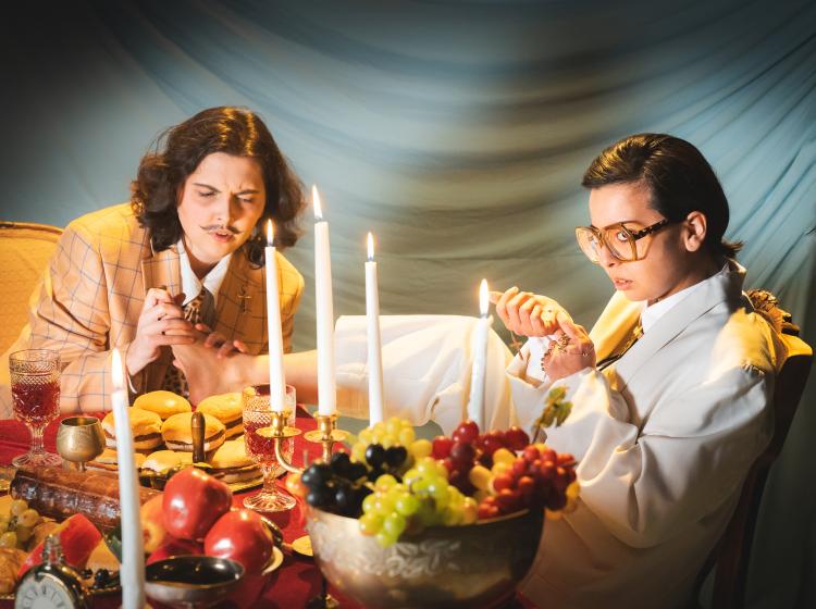 PSU Theater Arts' Tartuffe and Orgon sitting at the table laden with cheese burgers, fruit and candles. Tartuffe is looking at his gold cross with his bare foot on the table while Orgon massages the foot.