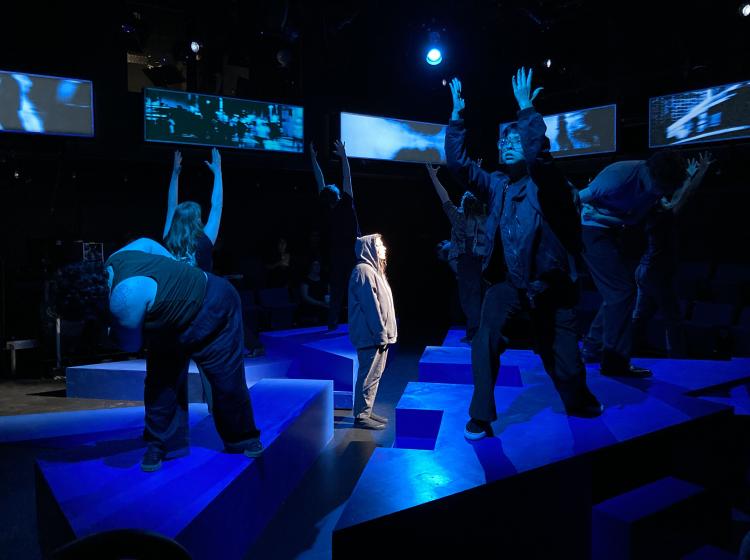 PSU cast of Caryl Churchill's "Love and Information" doing a dance interlude in the play on blue cubic stage blocks with one actor in the center lit with a spotlight.