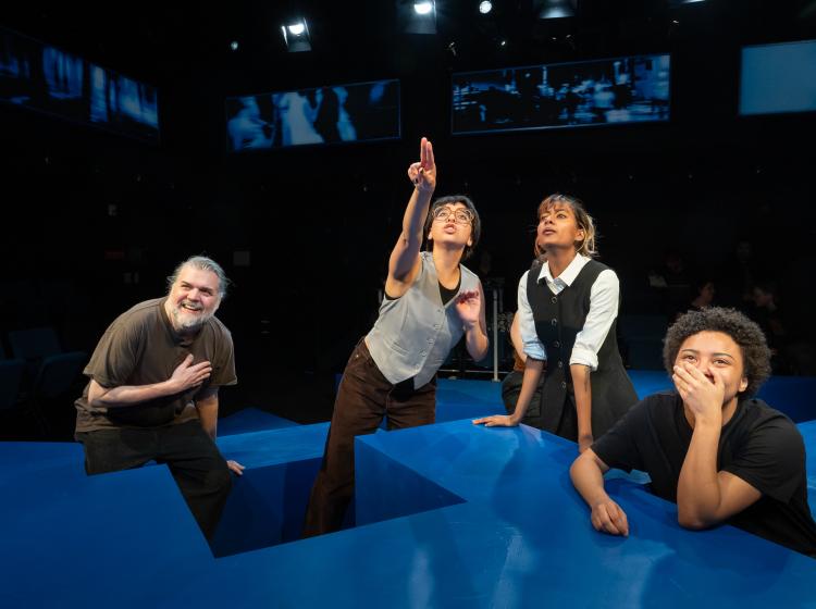 PSU cast perform in Caryl Churchill's "Love and Information." four people are looking up at a TV screen with varying reactions. One if pointing.