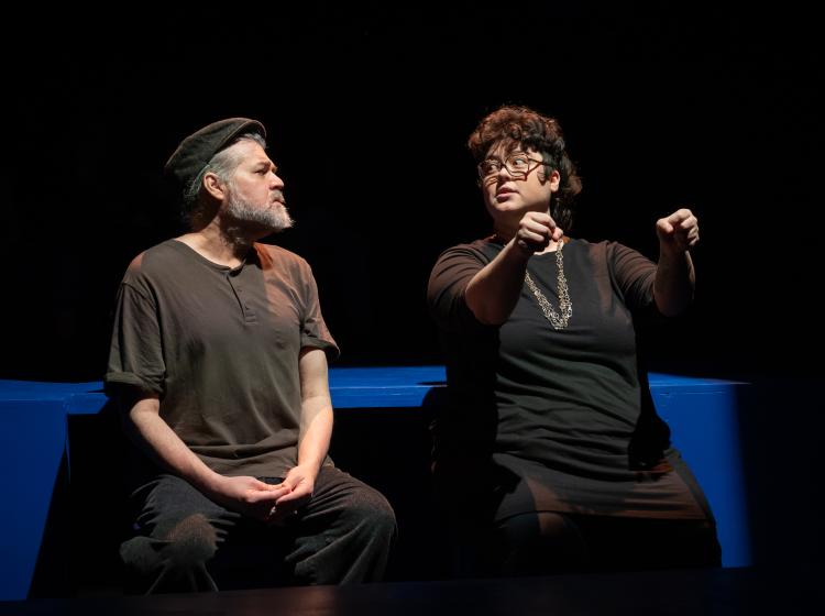 PSU cast perform in Caryl Churchill's "Love and Information." Two actors sitting on blue stage blocks are side by side looking at each other. One is holding an imaginary steering wheel.