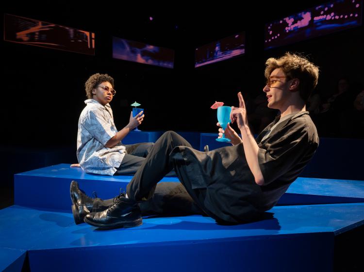 A scene from PSU Theater Arts production of "Love and Information." Two people lounge on blue stage blocks like they are lawn chairs, drinking tropical cocktails.