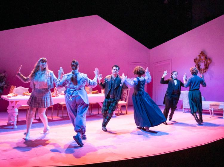 Six people in motion dancing in pairs with palms together turning on a bright pink stage.