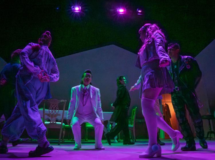 On a dark stage lit with dim pink and purple lighting, a man in a white suite is in a semi squatting position looking crazed, while other people walking in a circle around him.