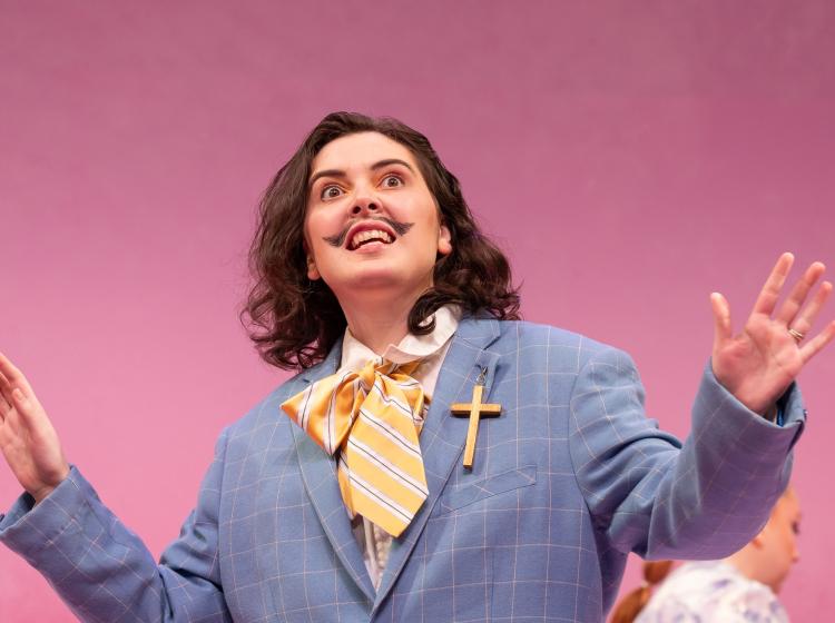 A man with long hair, a mustache, a dusty colored blue suit and a big cross on his label seems to be speaking and gesturing with his hands. There is a bright pink wall behind him.