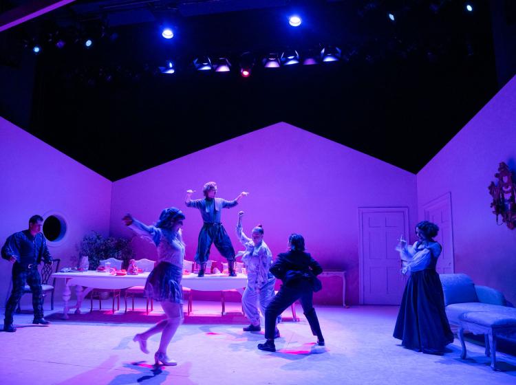 A dimly lit pink stage with a large dining table and five characters are dance partying. One of them is dancing on the dining table. Stage lighting can be seen above.