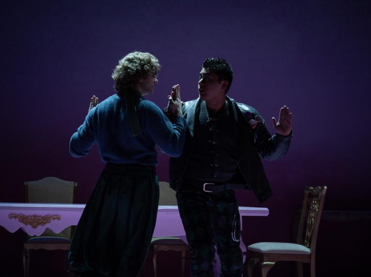 A dark stage with dim lighting. Two men seem to be dancing, palm to palm in a menacing stand off.