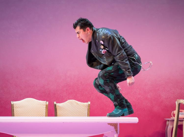 An actor wearing a black leather jacket, black boots, a chain at his hip, and with greased spiky hair, is in mid-motion of yelling and jumping on the end of a pink dining table, with a pink wall behind. 