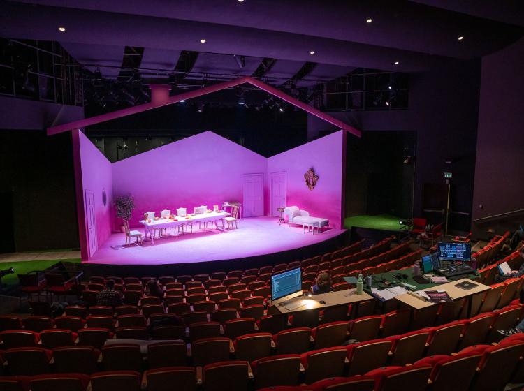 View from the top seats of a big auditorium, with the lighting and tech table nearby, looking down at the set of Tartuffe which is a large living room with three bright pink walls, a pink floor, the roofline of the house, a large dining table and a chaise lounge.